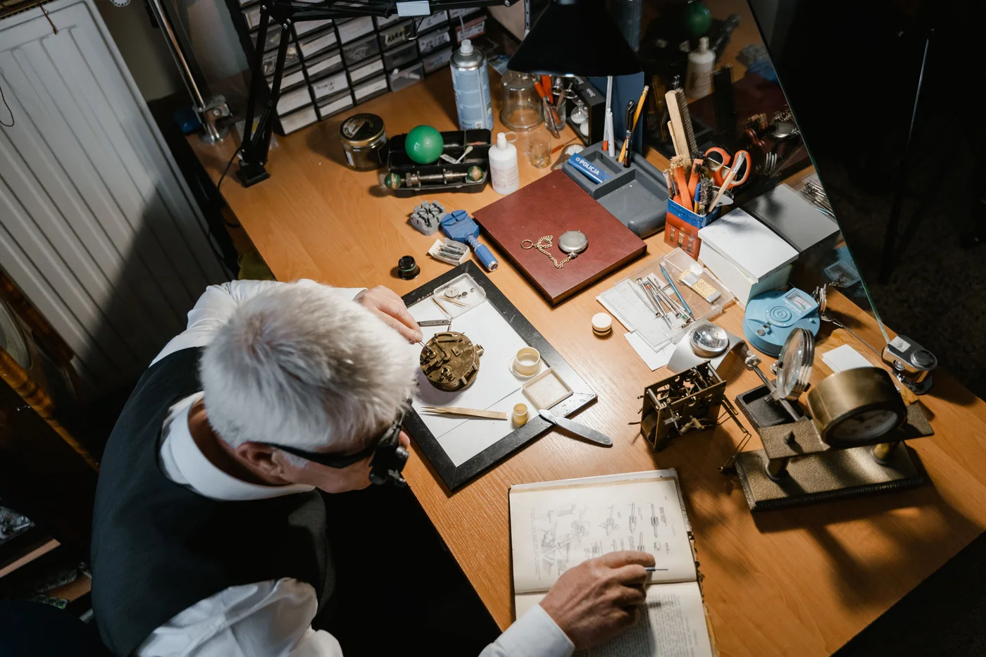 A master watchmaker at his bench, loupe to his eye, working on a movement with tweezers amid tools and an open reference book — the world of the independent watchmaker
