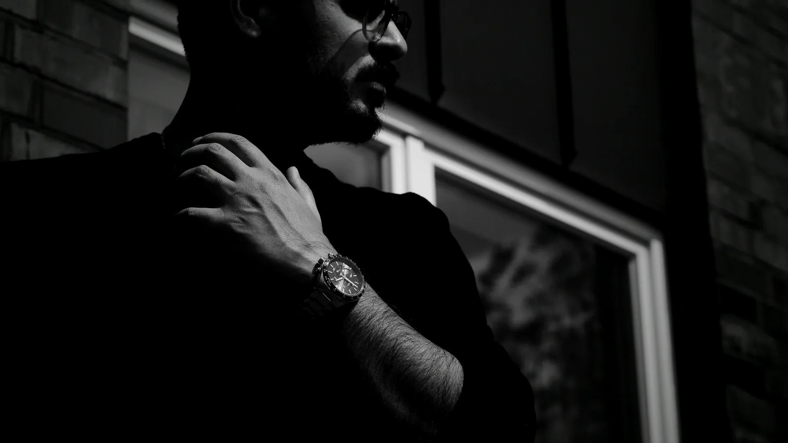 A man in profile wearing a chronograph wristwatch, hand at his collar — a collector and his watch, in moody black and white