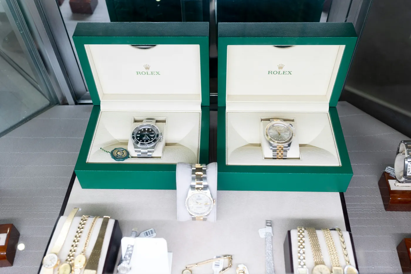 Two green Rolex boxes open on a dealer's display counter, each holding a watch, with the green Rolex certification seal beside one