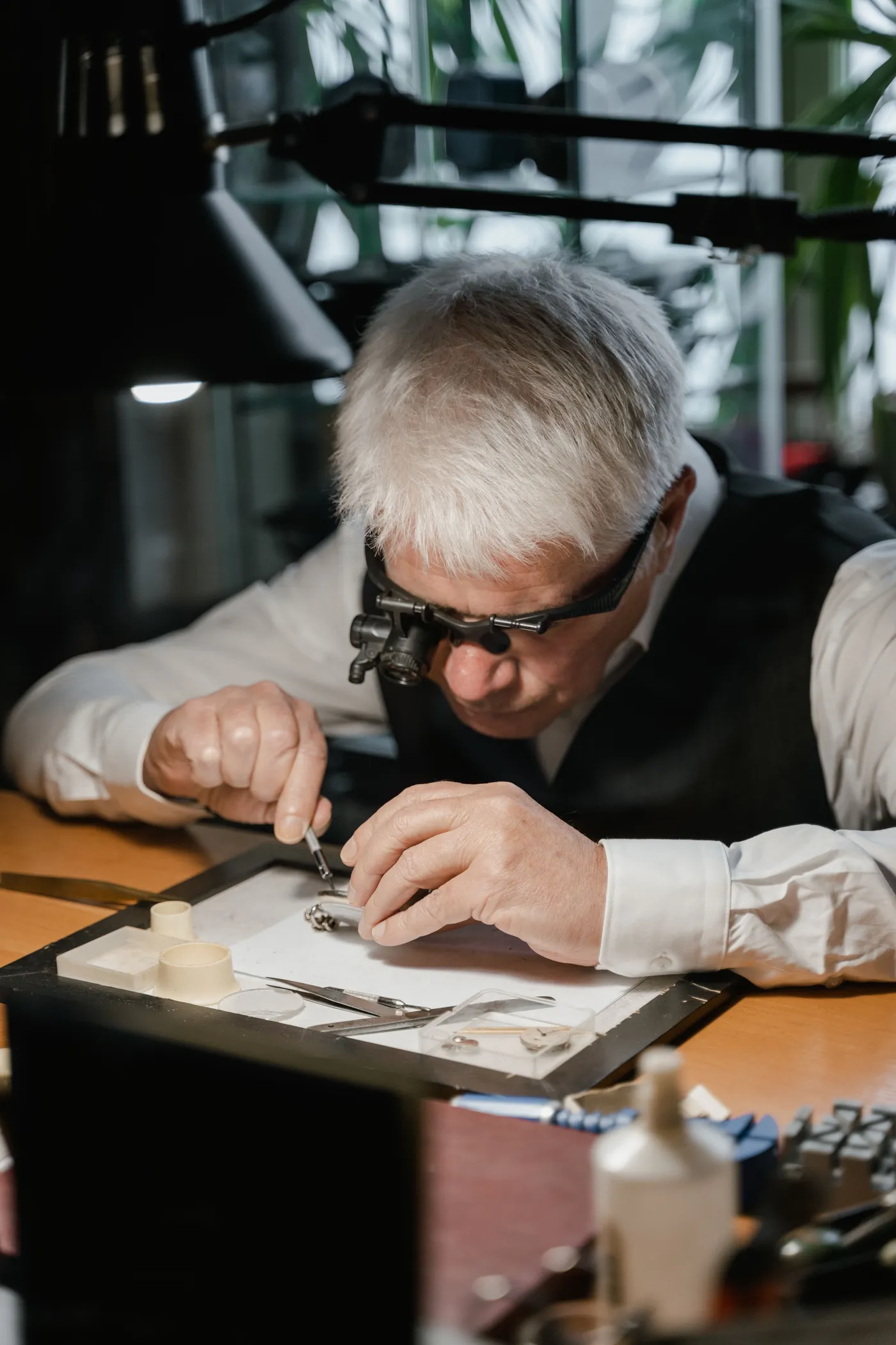 A watchmaker wearing a loupe inspects a watch component with tweezers at a workbench