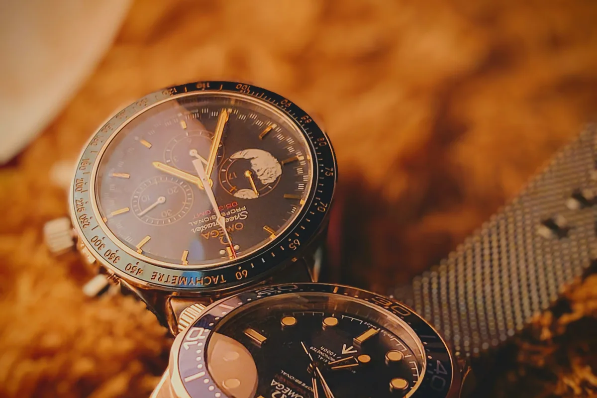 An Omega Speedmaster Professional Moonwatch chronograph with a moon-phase complication resting on a warm textured surface beside a second Omega diver