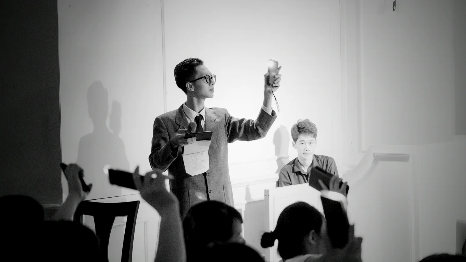A black-and-white auction-room scene — a presenter holds a lot up to the light as bidders in the foreground raise their phones