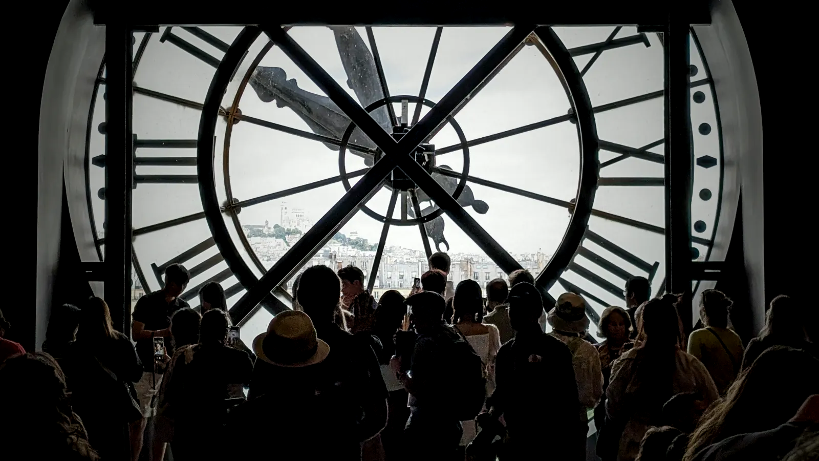 A crowd of visitors silhouetted against a giant backlit clock window, its Roman numerals and hands framing the city beyond